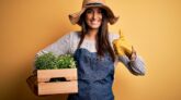 Young beautiful brunette gardener woman wearing apron and hat holding box with plants happy with big smile doing ok sign, thumb up with fingers, excellent sign