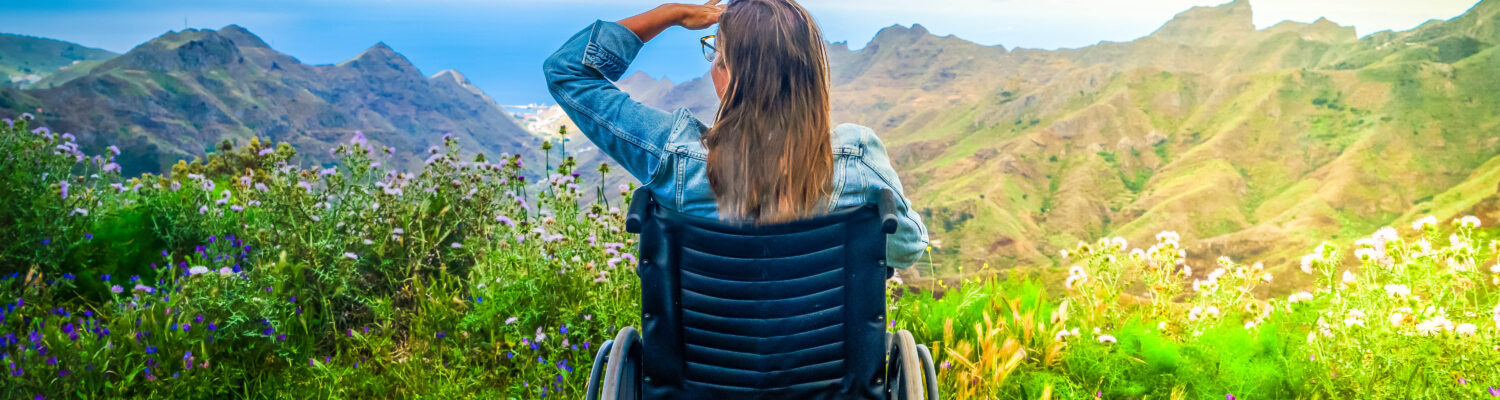 Never give up. Rear view of young handicapped woman sitting on wheelchair on top of mountain and looking at amazing nature landscape while traveling alone. International Disability Day