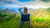 Never give up. Rear view of young handicapped woman sitting on wheelchair on top of mountain and looking at amazing nature landscape while traveling alone. International Disability Day