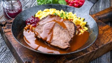 Traditional braised marinated German Sauerbraten from beef with spaetzle and cranberry jam in spicy brown sauce as closeup in a rustic wrought iron plate