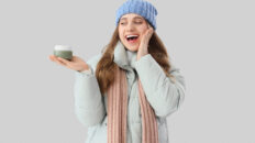 Happy woman in winter clothes and with cosmetic cream on grey background