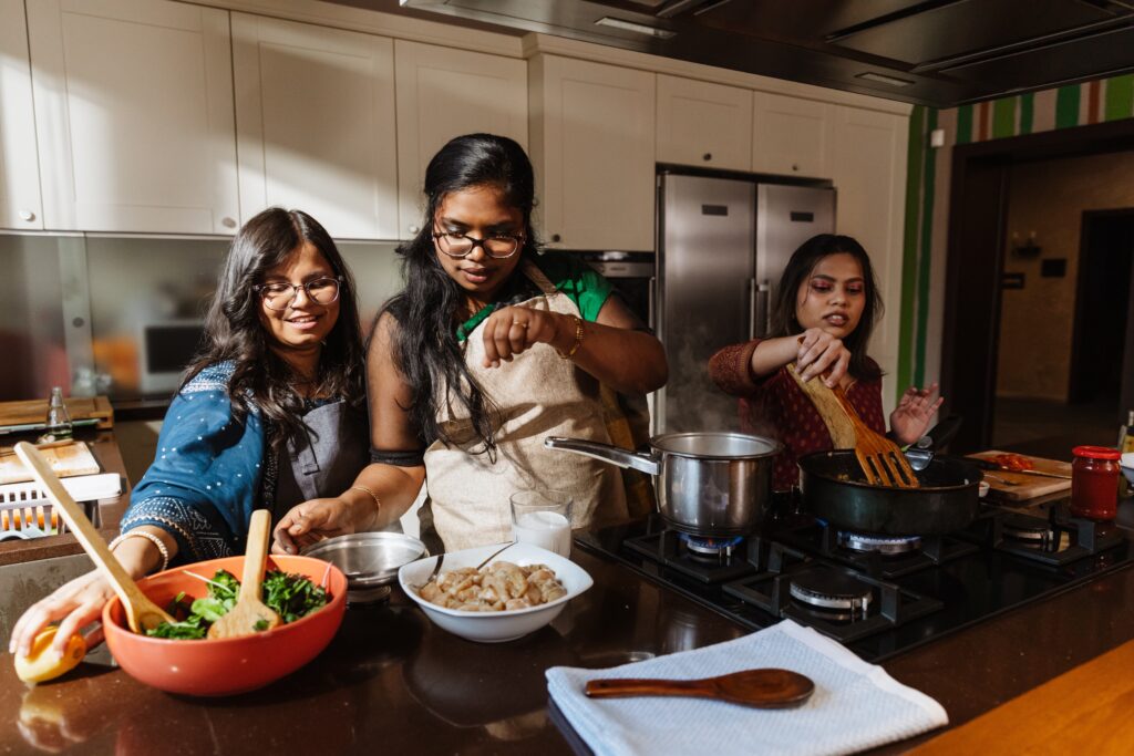 A woman stands and holds a spatula in a frying pan while a woman stands next to her, sprinkling spices, and a woman holding a lemon, in the kitchen