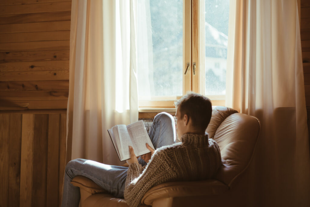 Young man in warm sweater reading book while relaxing on armchair by the window inside cozy log cabin