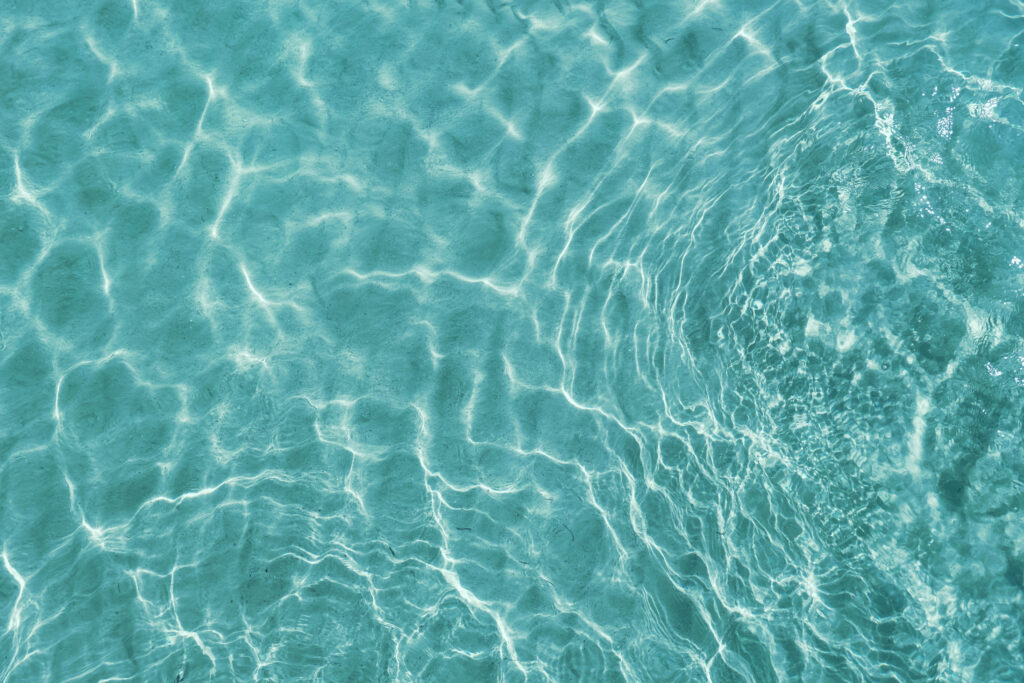 Water ripple over sandy beach. Transparent blue colored clear water surface texture with splashes. Water background, ripple and flow with waves. Blue water shinning. Sea, ocean surface. Overhead top view. Flat lay design.